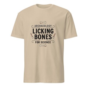 Peut inclure: Un t-shirt beige à manches courtes avec une impression graphique portant le texte ARCHAEOLOGY LICKING BONES FOR SCIENCE. Le motif comprend des illustrations de deux os et de deux petits pinceaux disposés autour du lettrage noir sur la poitrine.