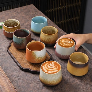 May include: A collection of eight ceramic cups with varied glaze finishes, including speckled blue, brown, orange, and cream tones. Two cups are filled with coffee featuring latte art. The cups are arranged on a rectangular woven tray.
