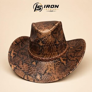 May include: A brown and black snakeskin patterned cowboy hat featuring a braided band around the base of the crown. The logo JS IRON STRIDE CO is printed in black and gold at the top centre above the hat against a solid light beige background.