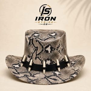 Peut inclure: Un chapeau à larges bords avec un motif peau de serpent gris et noir. Une bande en cuir noir entoure la calotte, ornée de cinq accents verticaux en forme de défense blanc cassé. Au-dessus du chapeau, le logo indique JS IRON STRIDE CO.