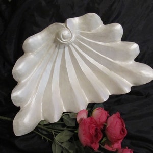 May include: A white ceramic seashell sculpture with a smooth, pearlescent finish. The shell is shaped like a fan with a raised center and delicate ridges. The sculpture is displayed on a black background.