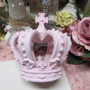 May include: A pink crown with a decorative design. The crown is made of ceramic and has a textured surface.