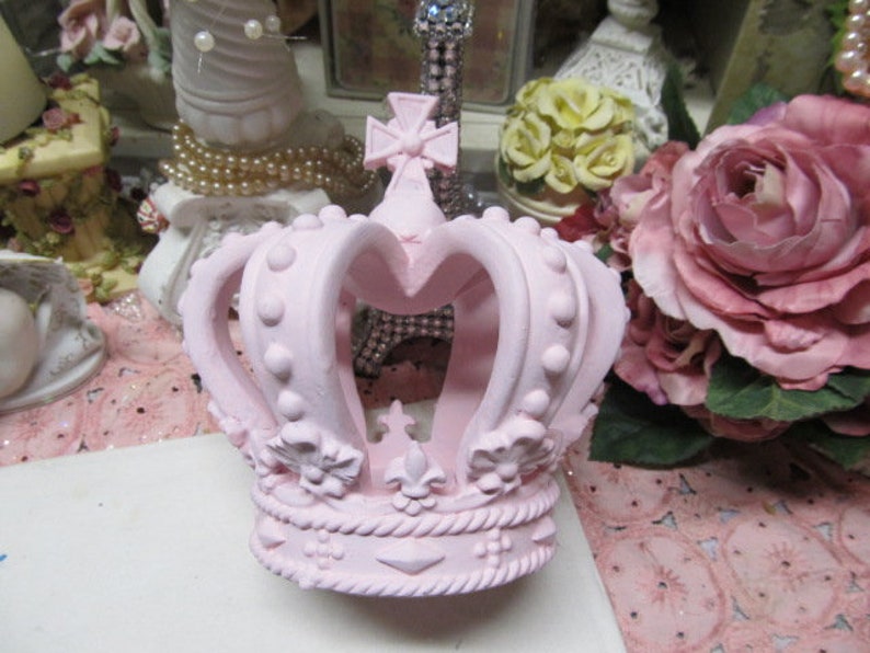 May include: A pink crown with a decorative design. The crown is made of ceramic and has a textured surface.