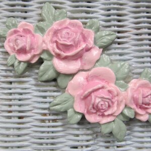 May include: A cluster of pink roses with green leaves, arranged in a decorative design. The roses are made of a smooth, textured material and have a realistic appearance.