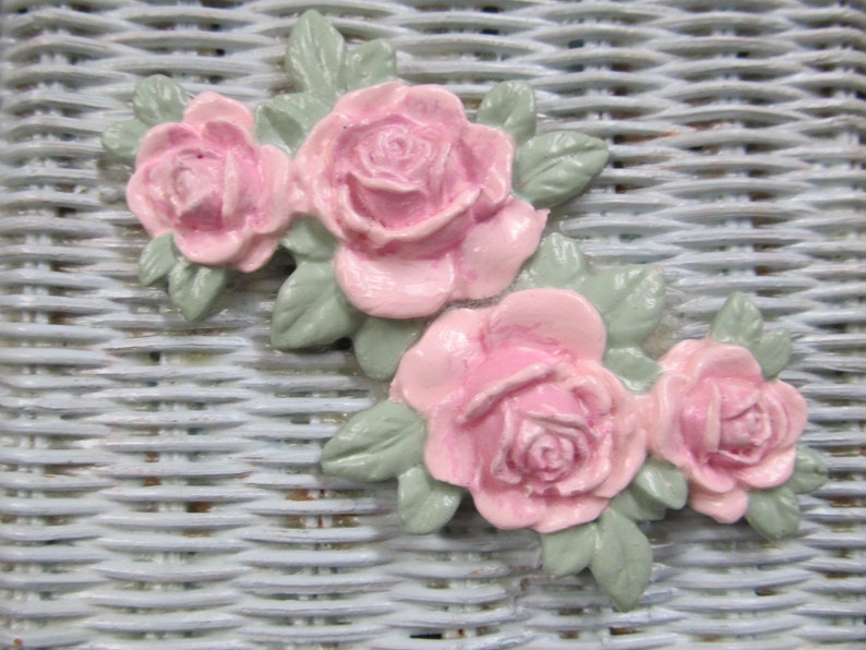 May include: A cluster of pink roses with green leaves, arranged in a decorative design. The roses are made of a smooth, textured material and have a realistic appearance.