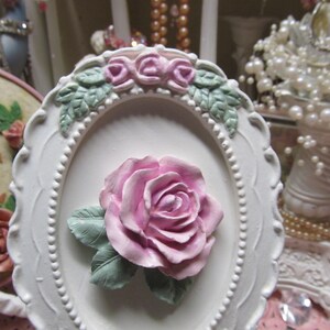 May include: White oval frame with a raised, pink rose and green leaves. The frame is decorated with a beaded border and a row of smaller pink roses.
