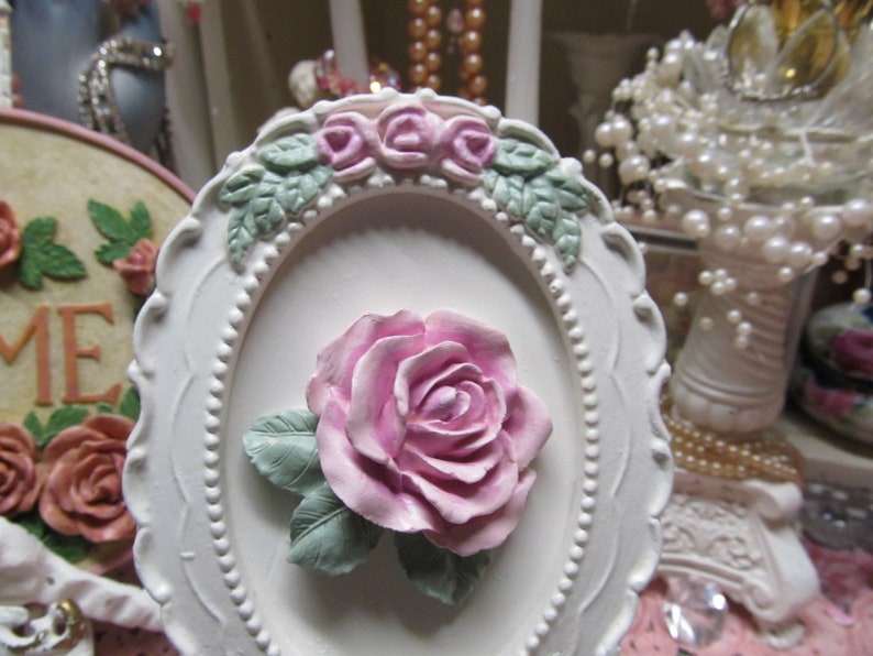 May include: White oval frame with a raised, pink rose and green leaves. The frame is decorated with a beaded border and a row of smaller pink roses.