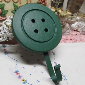 May include: A green wooden button-shaped hook with a metal hook. The button has four holes.