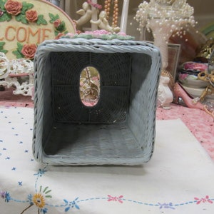 May include: A light blue wicker tissue box cover with a floral design. The cover has a square shape and an opening on the top for tissues.