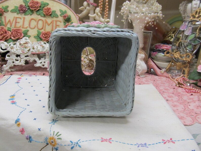 May include: A light blue wicker tissue box cover with a floral design. The cover has a square shape and an opening on the top for tissues.