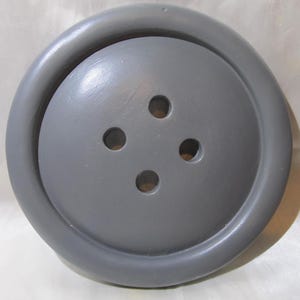 May include: A large, circular, grey button with a smooth, matte finish. The button features a raised outer rim and a slightly domed center section. Four evenly spaced, circular holes are arranged in a square pattern in the middle of the button.