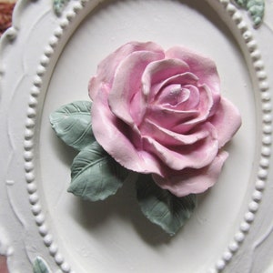 May include: A white oval frame with a raised, pink rose and green leaves. The frame has a beaded edge.