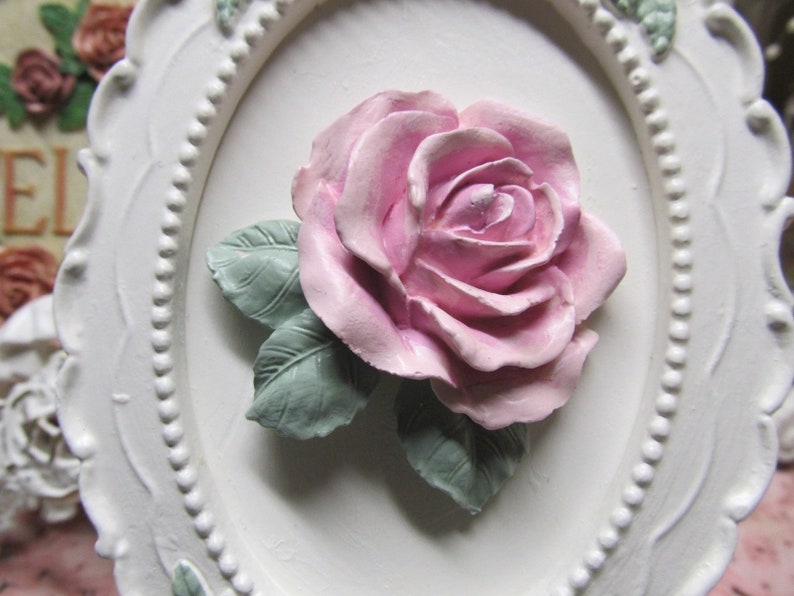 May include: A white oval frame with a raised, pink rose and green leaves. The frame has a beaded edge.