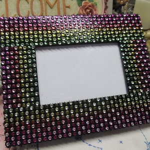 May include: A black picture frame with a rainbow pattern of rhinestones. The frame is made of black material and has a white mount.