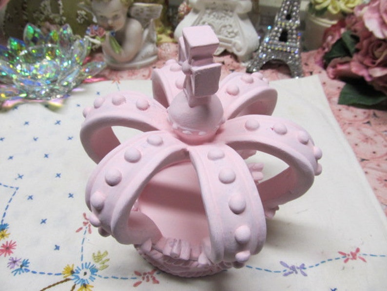 May include: A pink crown with a raised centre and a decorative design. The crown has a smooth, matte finish and is made of ceramic.