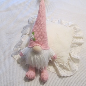 May include: A pink gnome with a white beard and a pink hat with a white flower. The gnome is sitting on a white pillow with lace trim.