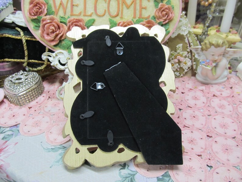 May include: Gold-colored ornate picture frame with a black velvet backing. The frame has a decorative design with floral motifs.