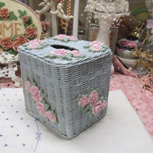 May include: A light blue wicker tissue box cover with pink roses and green leaves. The cover is woven and has a square shape.