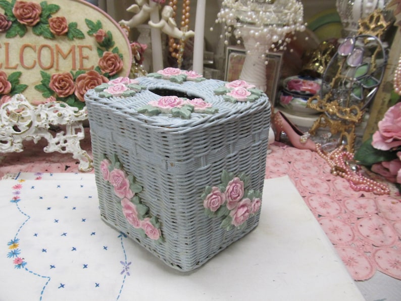 May include: A light blue wicker tissue box cover with pink roses and green leaves. The cover is woven and has a square shape.