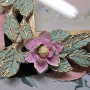 May include: A gold picture frame with a floral design. The frame has a pink flower with a yellow center and green leaves.