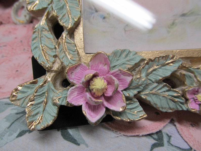 May include: A gold picture frame with a floral design. The frame has a pink flower with a yellow center and green leaves.
