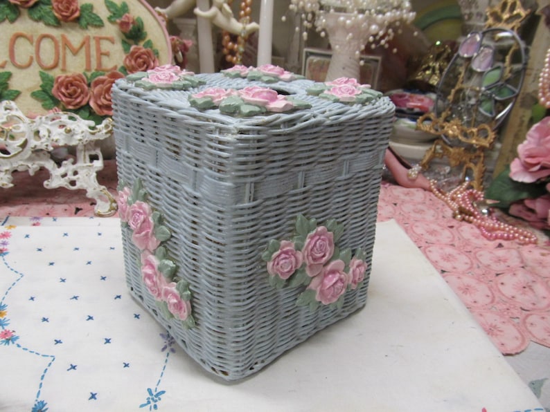 May include: A light blue wicker tissue box cover with pink roses and green leaves. The cover is square and has a woven design.