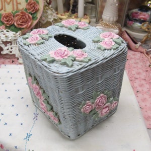 May include: A light blue wicker tissue box cover with pink roses and green leaves. The tissue box cover has a square shape and a cutout for tissues.