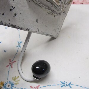 May include: White metal hook with a black knob on the end. The hook is attached to a white metal base with a decorative design.