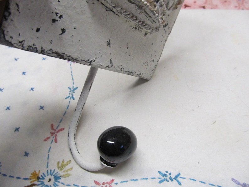 May include: White metal hook with a black knob on the end. The hook is attached to a white metal base with a decorative design.