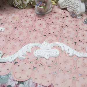 May include: White decorative architectural element with a distressed finish. The ornate design features scrolling details and a central flourish. The piece is set against a pink lace background, suggesting a vintage or shabby chic aesthetic.
