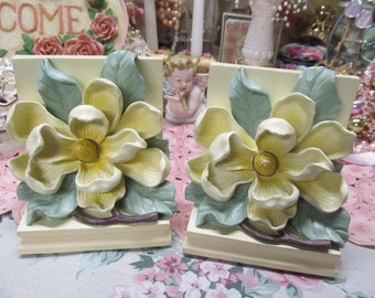 May include: A pair of bookends featuring a yellow magnolia flower with green leaves on a cream-colored base.