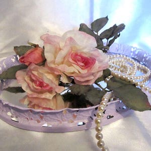 Pu&ograve; includere: Un vassoio in metallo color lavanda con un bordo decorativo a festoni, che presenta un bouquet di rose rosa e una collana di perle.