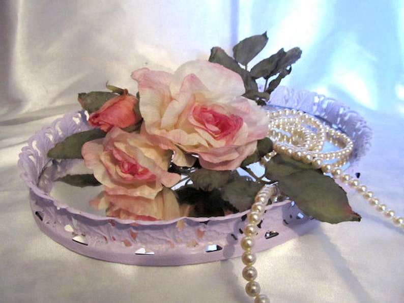 Pu&ograve; includere: Un vassoio in metallo color lavanda con un bordo decorativo a festoni, che presenta un bouquet di rose rosa e una collana di perle.