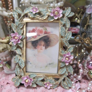 May include: A vintage-style picture frame with a gold finish and a floral design. The frame is decorated with pink flowers and green leaves. The frame is empty.