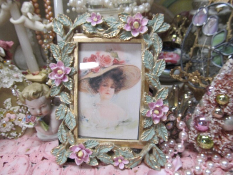 May include: A vintage-style picture frame with a gold finish and a floral design. The frame is decorated with pink flowers and green leaves. The frame is empty.