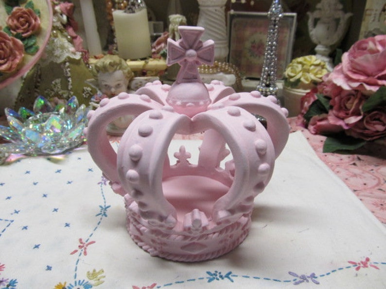May include: A pink ceramic crown with a cross on top. The crown has a raised design of dots and is perfect for a princess.