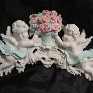 May include: A white decorative wall plaque featuring two cherubs with light blue drapes, holding a central basket filled with pink and blue flowers. The cherubs have detailed wings and are positioned symmetrically above ornate scrollwork carvings.