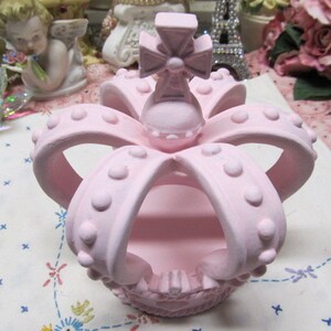 May include: A pink ceramic crown with a cross on top. The crown has a raised design of dots and is hollow in the centre.