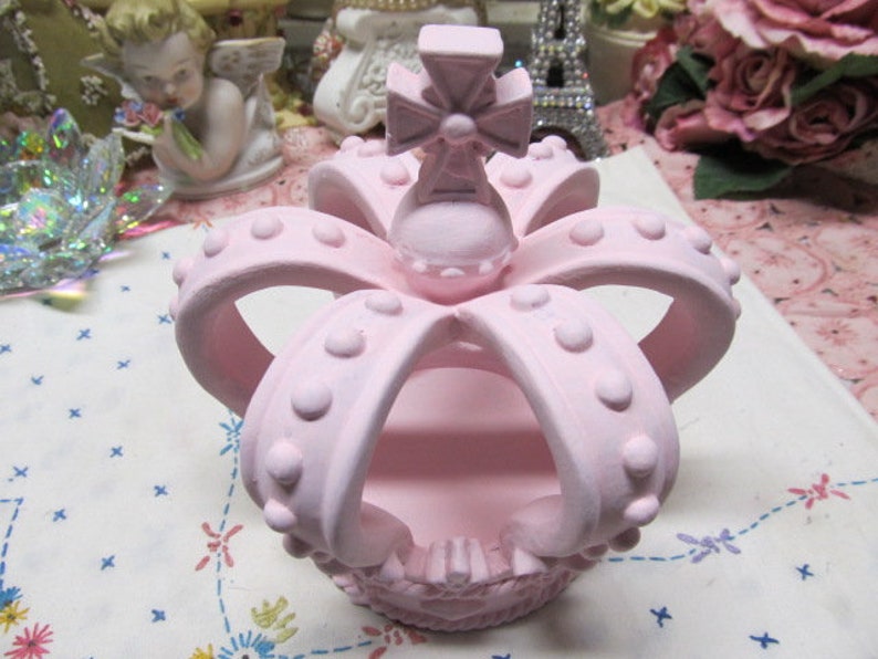 May include: A pink ceramic crown with a cross on top. The crown has a raised design of dots and is hollow in the centre.