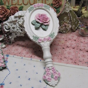 May include: White hand mirror with a pink rose design and a white handle. The mirror is surrounded by a white frame with pink roses and green leaves.