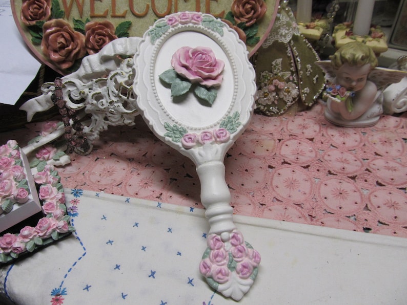 May include: White hand mirror with a pink rose design and a white handle. The mirror is surrounded by a white frame with pink roses and green leaves.
