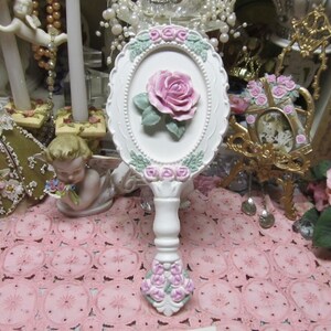 May include: White hand mirror with a pink rose and floral design. The mirror has a white handle and is surrounded by a white frame with pink flowers.