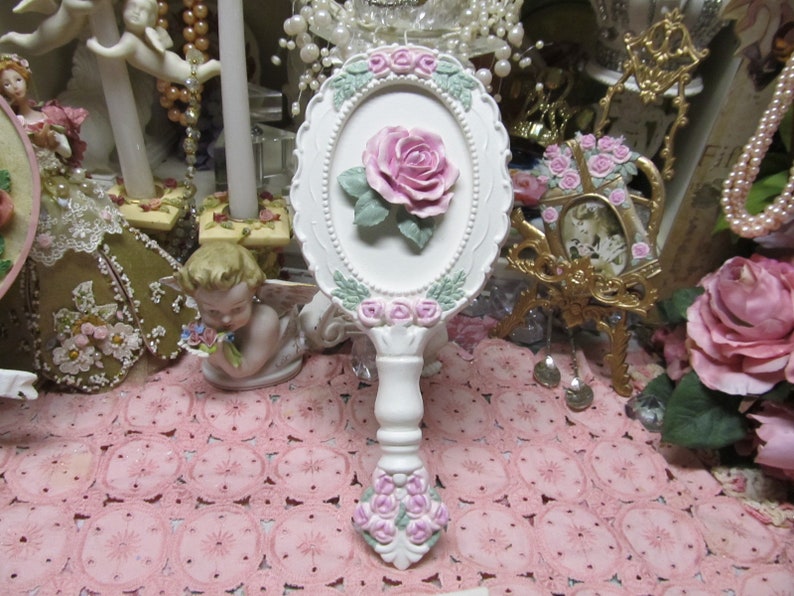 May include: White hand mirror with a pink rose and floral design. The mirror has a white handle and is surrounded by a white frame with pink flowers.