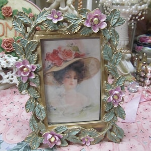 May include: A gold-toned picture frame with a floral design. The frame is decorated with pink flowers and green leaves. The frame is holding a vintage picture of a woman wearing a hat.