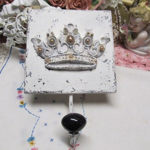 May include: White wooden square with a decorative gold and white crown design. A black knob hook is attached to the bottom of the square.