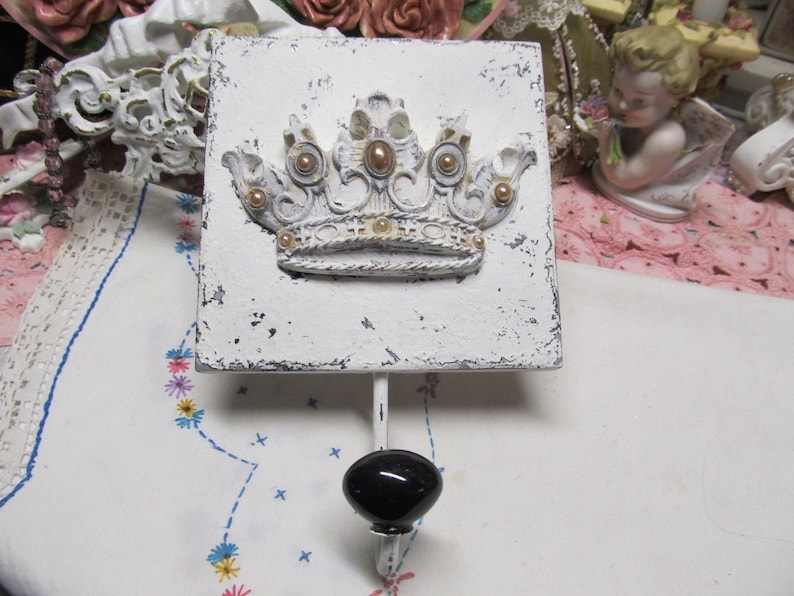 May include: White wooden square with a decorative gold and white crown design. A black knob hook is attached to the bottom of the square.