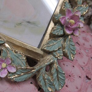 May include: A gold-toned picture frame with a floral design. The frame has a green and gold leaf design with pink flowers. The frame is on a pink surface.