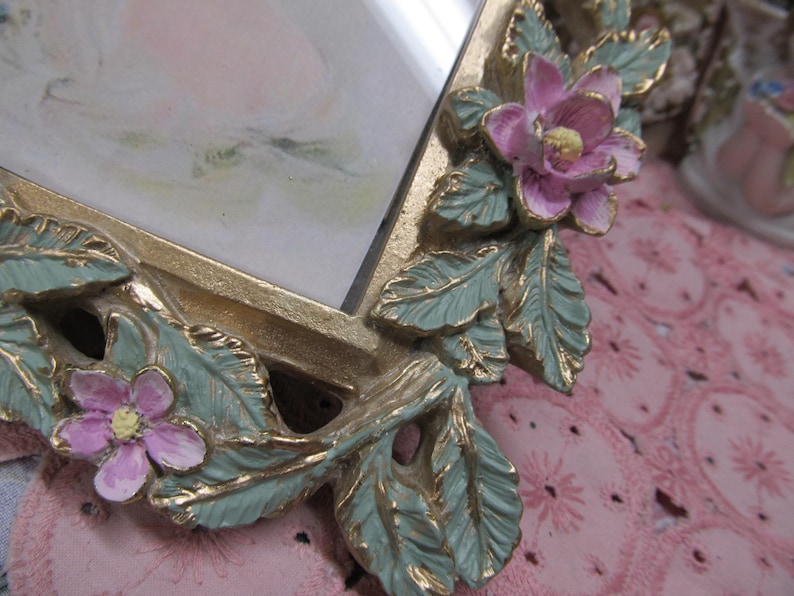 May include: A gold-toned picture frame with a floral design. The frame has a green and gold leaf design with pink flowers. The frame is on a pink surface.