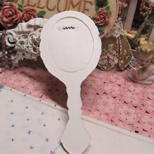 May include: White hand mirror with a decorative, ornate handle. The mirror is oval shaped and has a white painted finish.