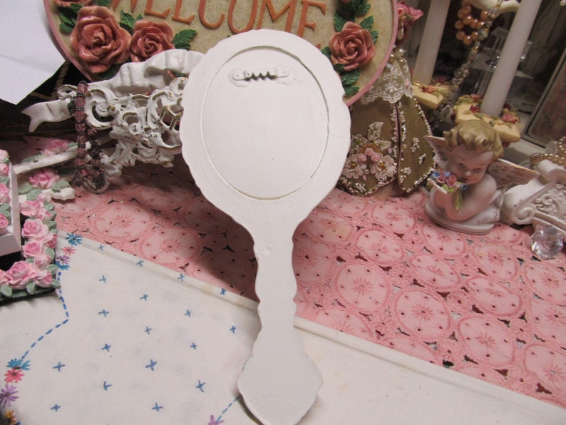 May include: White hand mirror with a decorative, ornate handle. The mirror is oval shaped and has a white painted finish.